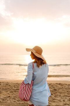 A sweet woman in a hat and carrying a red bag strolls along the beach at sunset. Young woman enjoys seascape in the sun's rays. Concept of relaxation, nature, and weekends.