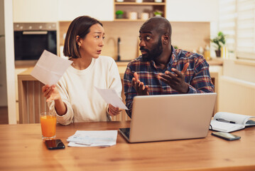Obraz premium A couple sits at a table reviewing bills and documents, looking concerned as they compare paperwork with information on a laptop.