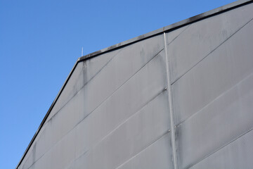 Fototapeta premium A grey corrugated metal facade of an industrial building with a diagonal roofline stands out against a clear solid blue sky background