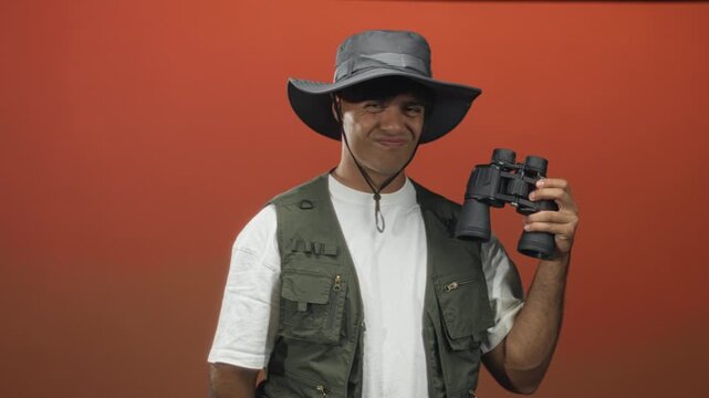Man wearing a wide brim hat and field vest holds binoculars and points a finger toward the viewer in a studio with an orange backdrop; curiosity.