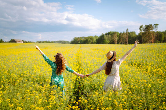 Two cheerful friends in dresses are having fun in a blooming field, bathed in sunlight. The young women pose outdoors, enjoying nature. Concept: style, fashion, and friendship.