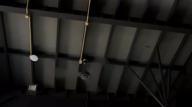 Dark industrial ceiling with hanging light fixtures. Metal beams and wires visible above. Light fixtures hang at different heights. Dark environment with low ambient light