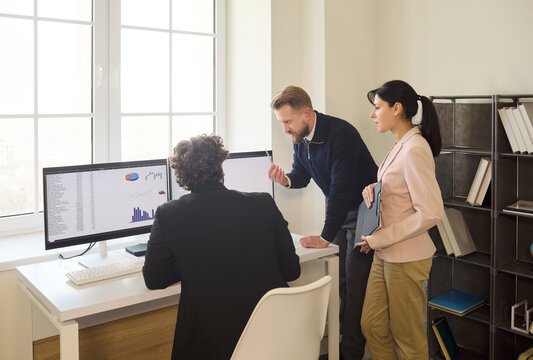 Team coworkers in meeting discuss office analysis. They review finance charts on dual computer monitors and align audit strategy in a bright workspace. Clear data driven teamwork concept.