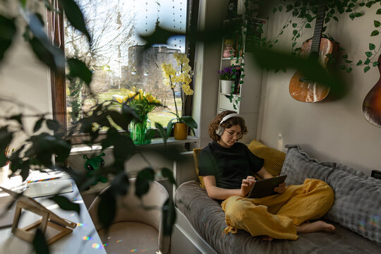 Young person with curly hair using tablet in cozy plant filled bedroom