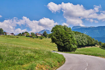 Obraz premium Road, countryside and farmland in nature with field, sky and clouds with mountains, trees and horizon. Outdoor, grass and buildings on hill with street, infrastructure or asphalt in springtime