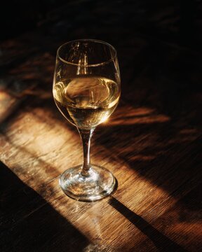 A single glass of white wine on a dark wooden table. A narrow beam of light illuminating the liquid and creating a caustic reflection.