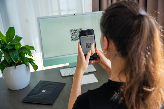 Person scanning QR code from computer screen with smartphone at home workspace. Digital interaction between devices and online service access concept.