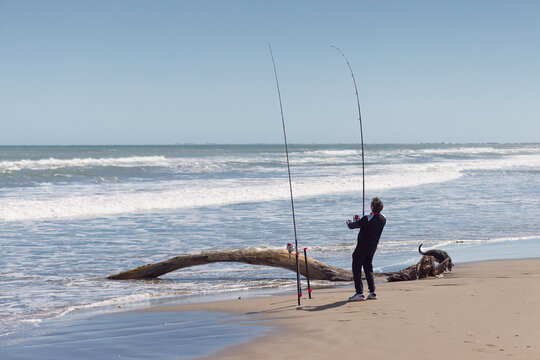 Man surf fishing with rods on ocean beach. Man standing on a sandy ocean beach next to two long surf fishing rods fixed in sand holders.