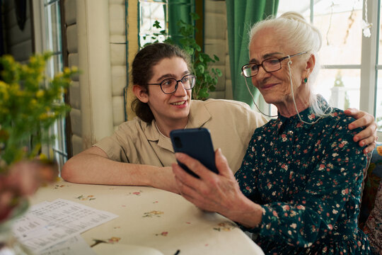 Senior Caucasian woman sitting at table holding smartphone while smiling teenager with long hair and glasses embracing her, both looking at phone screen and interacting together indoors