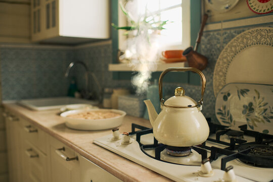 Vintage kettle heating on gas stove in cozy kitchen with sunlight streaming through window, ceramic dish with unbaked pie resting on countertop, blue patterned backsplash visible