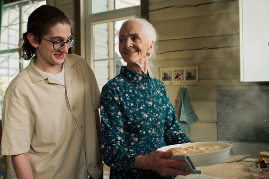 Senior Caucasian woman holding freshly baked pie while smiling at young adult Caucasian man standing beside her in kitchen, both sharing joyful moment during cooking process