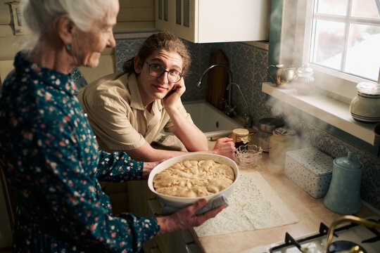 Senior Caucasian woman holding freshly baked pie while young Caucasian teenager with long hair sitting at kitchen counter looking at her, steam rising from pie