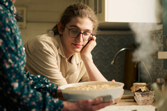 Caucasian teenager boy with long curly hair and glasses sitting at kitchen counter watching senior Caucasian woman holding freshly baked pie in dish, steam rising from food
