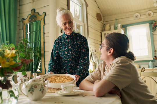 Senior Caucasian woman smiling while serving homemade pie to young adult Caucasian boy sitting at kitchen table, both engaging in warm interaction in rustic home setting