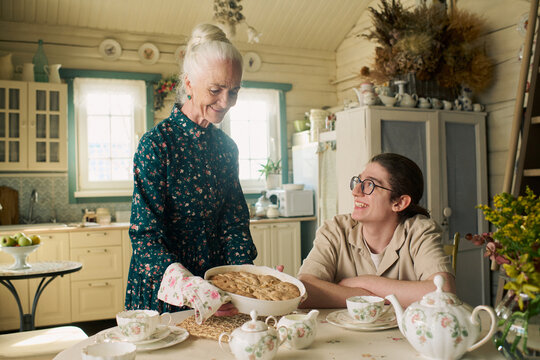 Senior Caucasian woman serving homemade pie to smiling teenage Caucasian boy sitting at table in cozy kitchen, both interacting warmly, surrounded by tea set and rustic decor