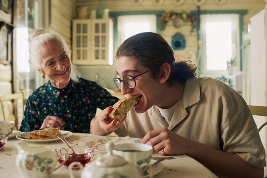 Senior Caucasian woman smiling while watching teenage Caucasian boy eating pie at dining table, both sitting in cozy kitchen setting with tea cups and plates visible