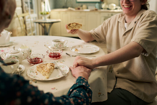Young Caucasian man smiling while holding elderly Caucasian womans hand and offering slice of pie at table set with tea and desserts, both sitting together in kitchen