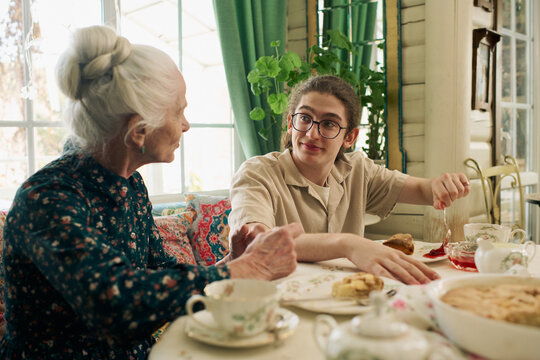 Senior Caucasian woman talking with teenage Caucasian boy sitting at table, teenager smiling and adding jam to pastry, both engaging in conversation during breakfast or tea time