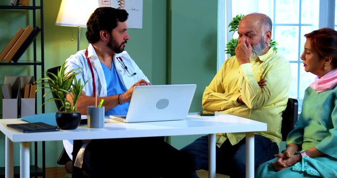 Professional Indian young male doctor using laptop computer with Asian senior retired couple patients across a table in a modern clinic; healthcare, digital technology, and old age care concept.