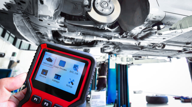 Car Diagnostic Scanner Tool, A technician holds a red diagnostic OBD2 scanner with a digital screen, showing menu icons, in a workshop while a car is on a lift.
