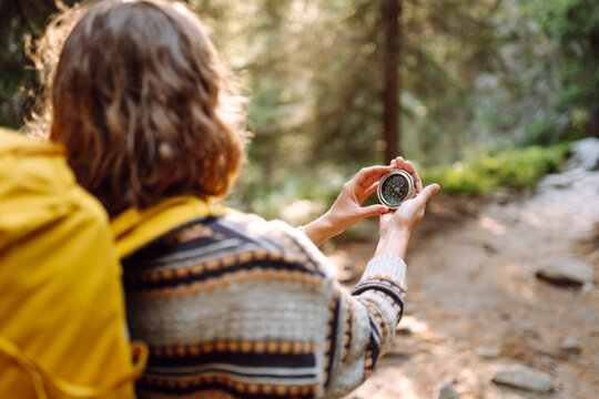 Rear view of young woman with hiking backpack holding compass while standing on hiking trail in mountains. Woman holds compass and looks for directions while hiking. Navigation concept, hiking.