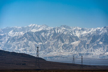 Obraz premium Snow covered Qilian Mountains in China with power lines in the foreground under a clear blue sky Snow covered mountains rise majestically behind a rugged landscape with power lines 