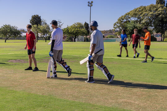 Male teammates in shorts and shirts walking across pitch at park carrying cricket bats and pads
