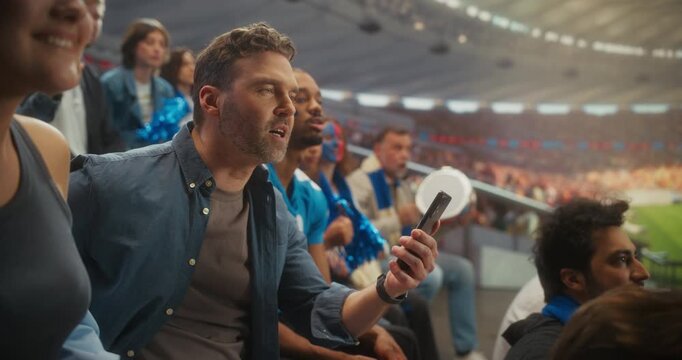 Stadium Championship Match: Man Using Smartphone for Online Sports Betting. Fan Checks Odds During Play, Watching the Team Win. Great Reaction as the Crowd Cheers Around