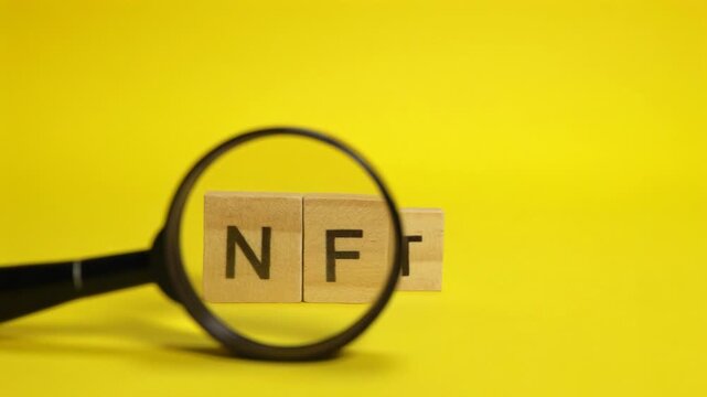 Magnifying glass moving over wooden cubes with the acronym nft on a bright yellow background