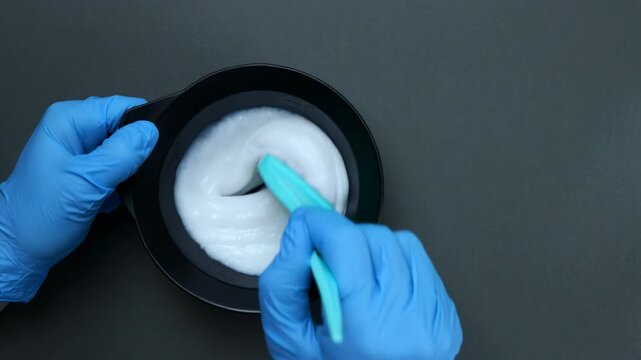 Professional hairdresser in blue gloves mixing hair color cream in a black bowl on a dark surface