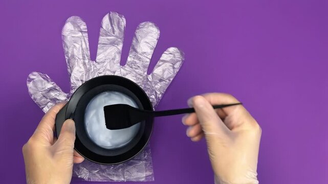 Professional hairdresser with gloves mixing hair dye in a plastic bowl on a purple background