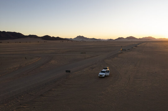 Aerial view of a lone white vehicle stands out against the stark, sandy landscape under the vast sky, as the sun rises over the distant mountains, Windhoek, Namibia.