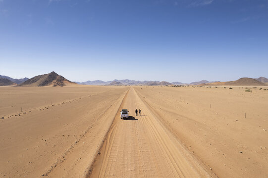 Aerial view of a lone white vehicle and two figures venture along a sandy track stretching into the horizon, framed by arid landscapes, Namibia.