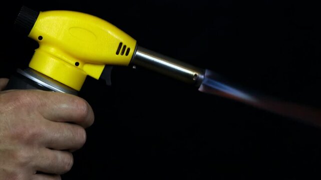 Man holding a lit yellow propane blowtorch with a blue flame on a black background