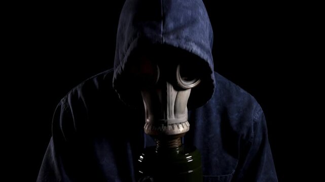 Mysterious figure in a dark hoodie and gas mask slowly bowing their head on a black background