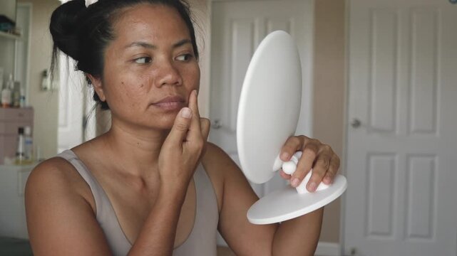 Young woman examines acne using a handheld mirror at home, touching her chin and observing skin condition in a calm, natural setting.