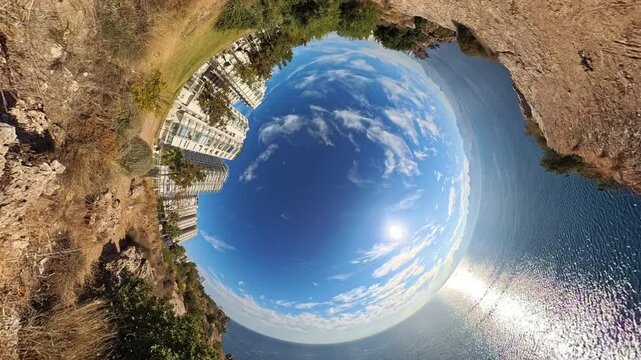 Tiny planet effect of a coastal area with buildings, greenery, and a view of the sea, creating a surreal, circular perspective of the landscape