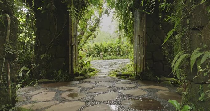 Ancient stone gate draped in moss and ferns opens pebbled path, inviting to serene, secret tropical garden in dense jungle foliage. Travel to Indonesia, explore Bali culture. Natural background
