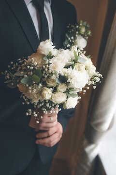 A groom holding a bridal bouquet of white roses with ribbon, preparing for an outdoor wedding ceremony. elegant black suit, formal attire, romantic mood