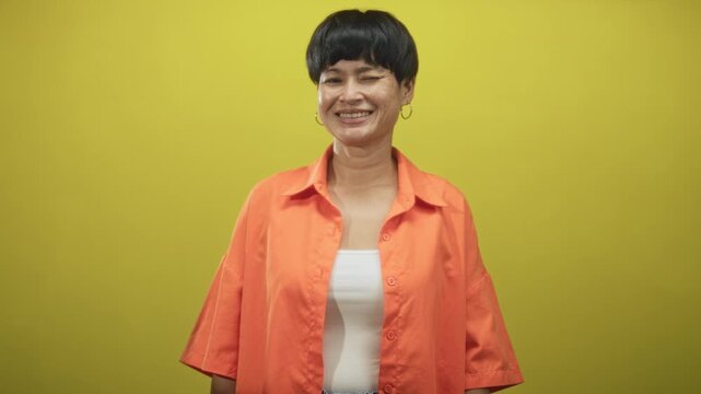 Woman winks and smiles, showing left eye and hoop earring, wearing orange shirt over white tank and short haircut against yellow studio backdrop; playful charm.