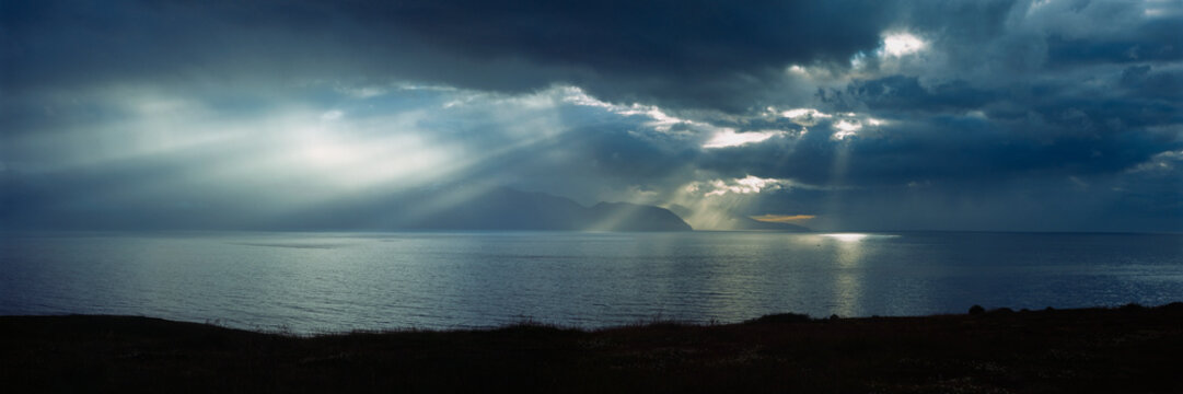 Panoramic view of sunbeams cutting through thick, dark clouds over tranquil blue lake, Iceland
