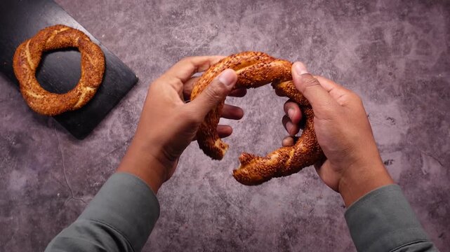 Person breaks Turkish bagel in half with sesame seeds on a textured surface.