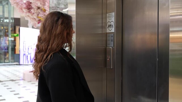 Side camera view of woman pressing elevator call button and waiting near stainless steel lift doors in modern shopping mall interior. Urban mobility, accessibility and daily routine