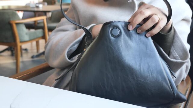 Woman's hands searching inside black leather handbag with strap in cafe.