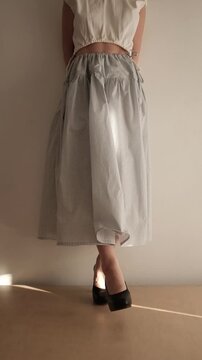A young woman with a brunette bob hairstyle showcases a stylish two-piece outfit featuring a white open-back crop top and a high-waisted, tiered gingham check maxi skirt in a minimalist indoor setting