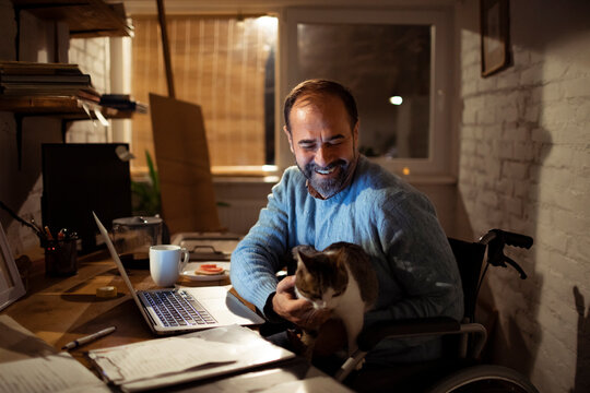 Smiling man in wheelchair with cat working on laptop at home office at night