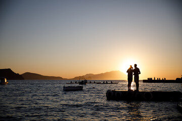 Fototapeta premium Wide sunset seascape with two silhouetted people standing on a floating dock, small boats and distant paddlers on the water.