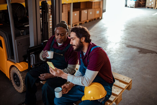 Two warehouse workers on coffee break using smartphone by forklift