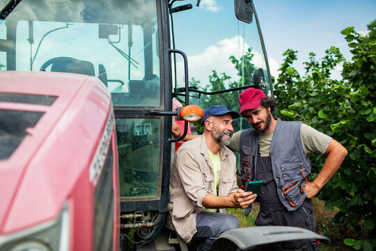 Two farmers using smartphone beside tractor in orchard