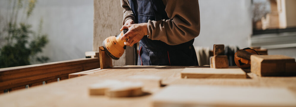 A carpenter uses an orbital sander to smooth a wooden plank on a workbench. The close-up scene shows hands operating a power tool in a workshop setting.